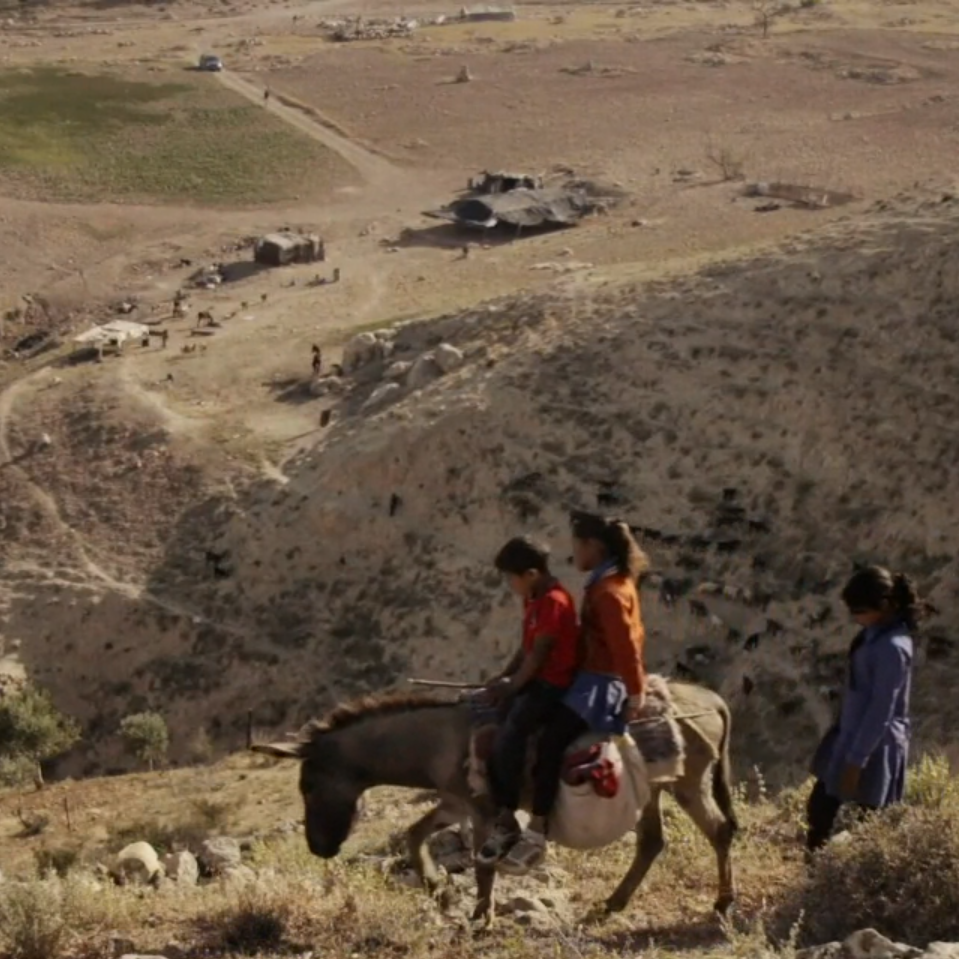 Ein Mädchen und ein Junge reiten auf einem Esel in einer trockenen Landschaft. Ein Mädchen läuft hinterher.