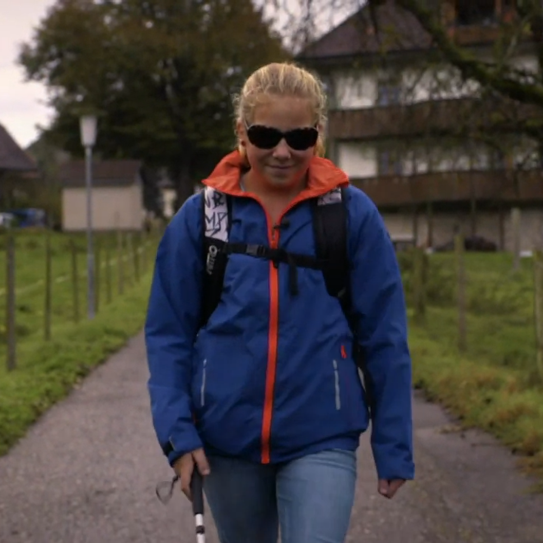 Ein Mädchen mit einer dunklen Sonnenbrille und einem Blindenstock geht lächelnd auf einem Weg.